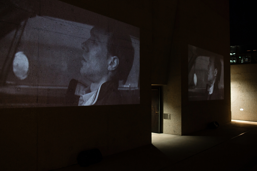 Fotografie einer Doppelvideoprojektion des Films "Mittwoch" des Künstlerduos M+M auf der Außenfassade des Eingangstors des Staatlichen Museums Ägyptischer Kunst. Auf den gezeigten Schwarz-Weiß-Videostills ist der gleiche Mann in Profilansicht zu sehen, der in der Dunkelheit in einem Auto fährt. Seine Miene ist ernst.