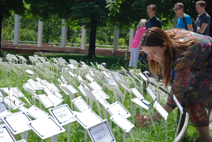 Photograph of a lawn in the Hofgarten with many white signs stuck into it. Words and sentences are written on the signs in different colours. A woman can be seen sticking another sign into the lawn. A few passers-by on the footpath and the Bavarian State Chancellery can be seen in the background.