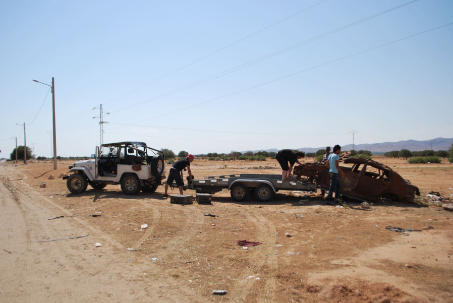 Picture of a burnt-out car wreck on the side of a gravel road in Tunisia. Four men are loading the wrecked car onto a trailer pulled by a white SUV.