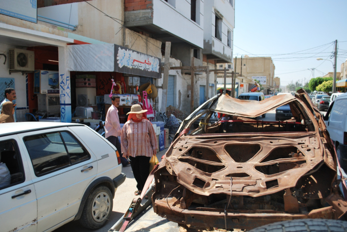 Image of a burnt out brown car wreck strapped to a car trailer and being towed through the streets of Sidi Bouzid, Tunisia. Parked and passing cars can be seen alongside the wrecked car, as well as passers-by watching the scene.