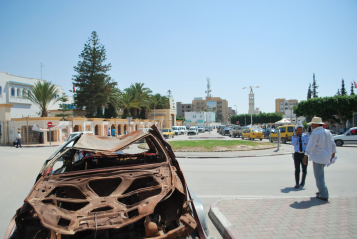 Image of a burnt out brown car wreck on the side of the road in Sidi Bouzid, Tunisia. In the background is a busy road with a roundabout and taller houses.