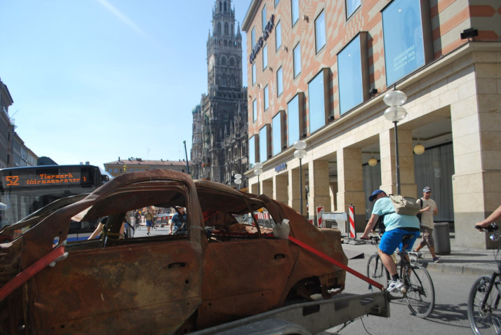 Photograph of a brown burnt-out car wreck without tyres on Munich's Marienplatz in front of Ludwig Beck. The wreck is secured to a trailer with red lashing straps. A city bus on route 52 can be seen in the background. Cyclists ride past the side of the car trailer and watch the unusual goings-on.