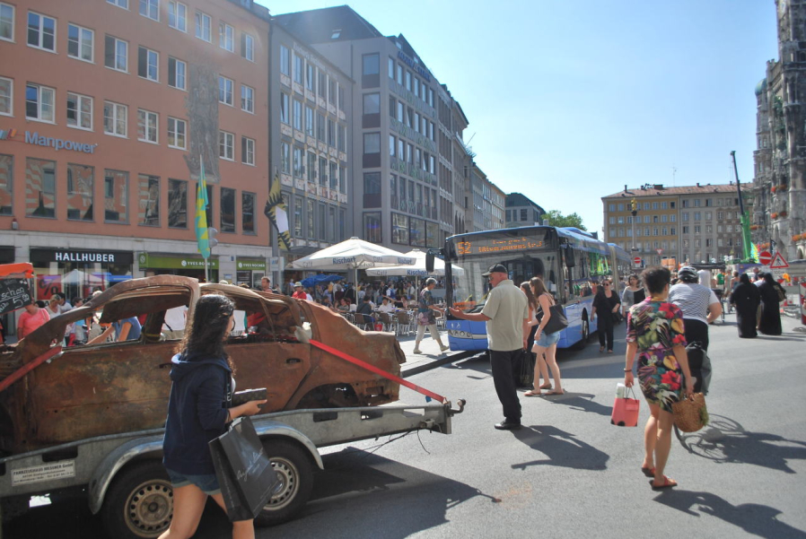 Photograph of a brown, burnt-out car wreck with no tyres in Munich's Marienplatz. The wreck is secured to a trailer with red lashing straps. In the background is a city bus on route 52. Passers-by in the square watch the unusual scene.