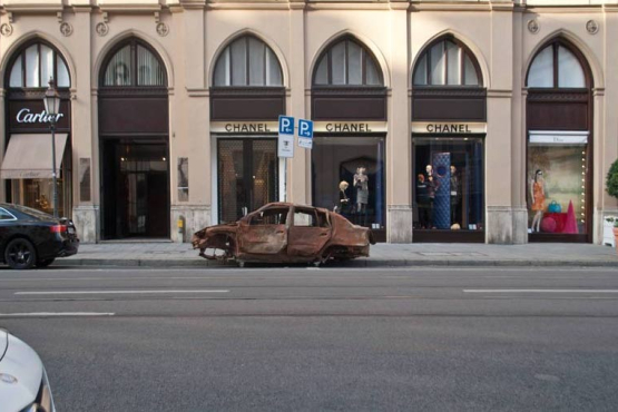 Photograph of a brown burnt-out car wreck without tyres parked on Maximilianstraße in Munich in front of the Chanel shop.