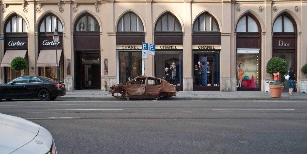 Photograph of a brown burnt-out car wreck without tyres parked on Maximilianstraße in Munich in front of the Chanel shop.