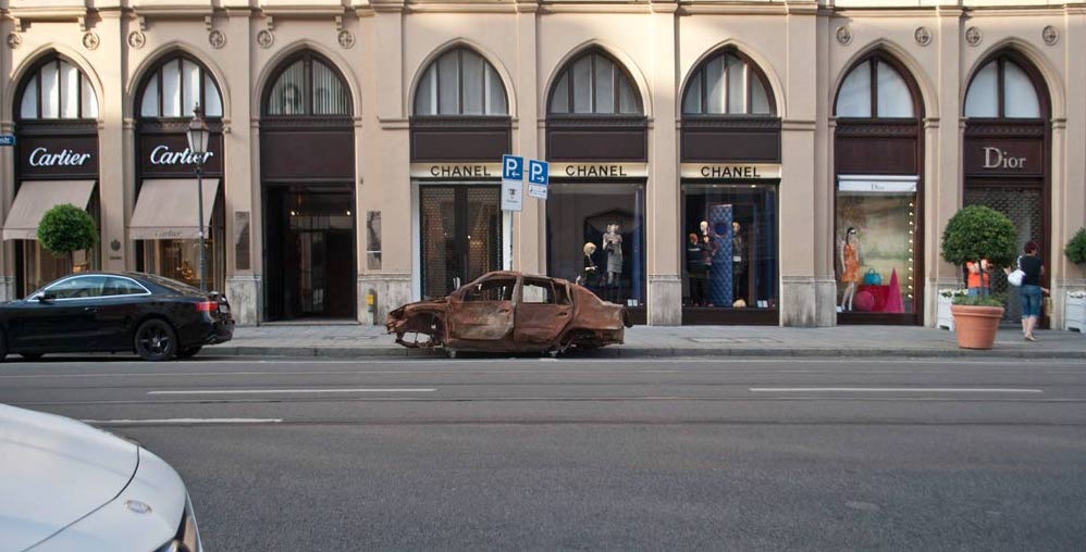 Photograph of a brown burnt-out car wreck without tyres parked on Maximilianstraße in Munich in front of the Chanel shop.