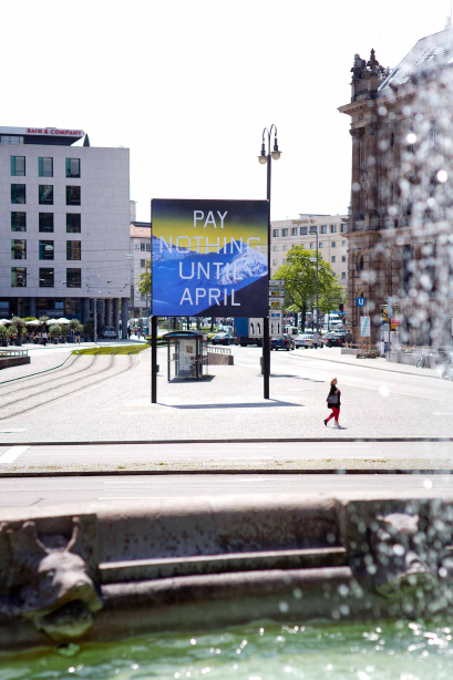 View of Lenbachplatz towards the city centre, taken from the Wittelsbacher Fountain. The edge of the fountain and the basin filled with water can be seen in the foreground. In the background is Lenbachplatz, with the billboard in the centre of the square. The billboard features a work by the artist Ed Ruscha. It shows a snow-covered mountain range in blue with a yellowish horizon above. The words "Pay Nothing Until April" are written in white capital letters on the image.