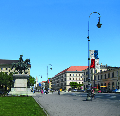 View of Ludwigstraße with pedestrians and cyclists seen from Odeonsplatz. As part of an art installation by artist Silke Witzsch, the national flags of various countries are attached to the street lamps at half height. The flags are artistically distorted by placing a camouflage pattern over the national flags.