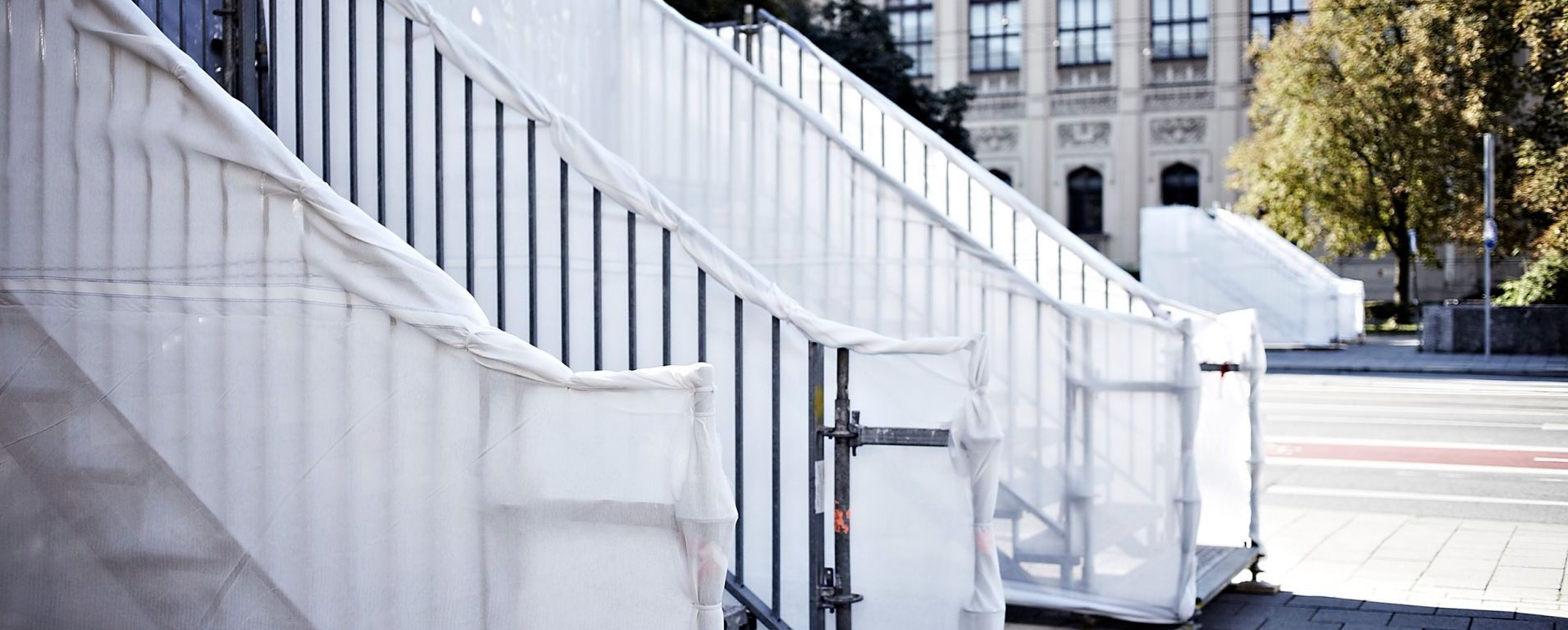 Die Fotografie zeigt zwei tribünenartige Stahltreppen, die mit weißem Stoff ummantelt sind. Die Tribünen sind auf beiden Seiten der Maximilianstraße jeweils vor den Unterführungsaufgängen, die nun zum MaximiliansForum führen, aufgestellt. Im Hintergrund zu erkennen, ist das Gebäude des Museum Fünf Kontintente, in der Bildmitte führt die Maximilianstraße entlang.