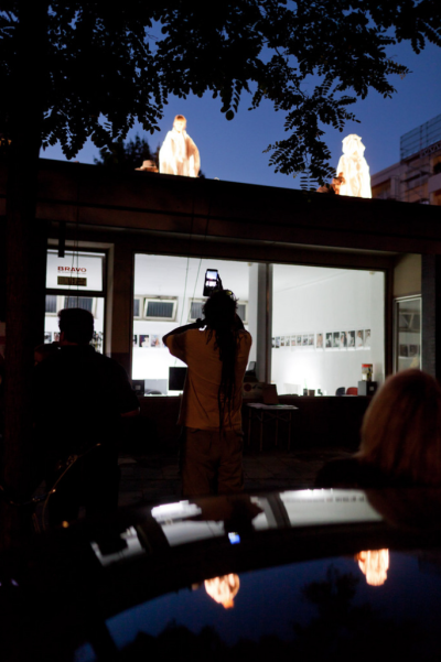 Photograph of a nocturnal performance on a single-storey building. The interior of the building is illuminated in a whitish colour and features office equipment and a row of small-format photographs on the walls. Two performers stand on the roof of the building, their white costumes seeming to glow from within.