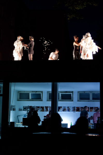 Photograph of a nocturnal performance on a single-storey building. The bluish-lit interior of the building shows office equipment and a row of small-format photographs on the walls. Some people are standing inside the building. Two female performers are standing on the roof of the building, their white costumes being adjusted by two black-clad dressing assistants. Another woman kneels in the centre of the roof and appears to be setting the scene.