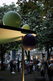 Photograph of the single-storey tram shelter on Habsburgerplatz. An installation of spherical objects of different sizes in green and blue is mounted on the protruding roof of the shelter. Surrounding the shelter are groups of people chatting.