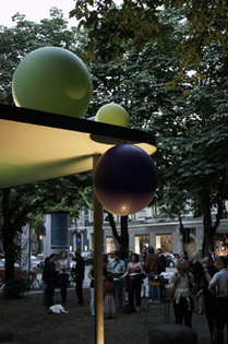 Fotografie des einstöckigen Tram-Wartehäuschens am Habsburgerplatz. Am vorspringenden Dach des Häuschens ist eine Installation aus unterschiedlich großen kugelförmigen Objekten in grün und blau angebracht. Um das Häuschen herum stehen Gruppen von sich unterhaltenden Mensch.