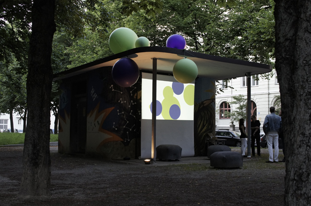 Photograph of the single-storey tram shelter on Habsburgerplatz. An installation consisting of five ball-shaped objects of different sizes in green and blue is mounted on the protruding roof of the shelter. Three grey poufs are placed under the roof. A video is also projected onto the roofed front of the house. It shows the motifs of the green and blue balls on a white background.