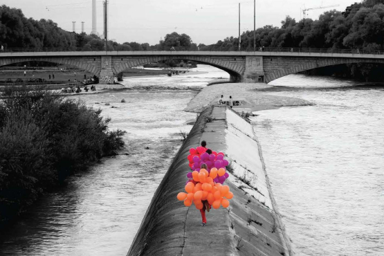 Black and white photograph of the Cornelius Bridge looking south towards the Reichenbach Bridge. On a concrete embankment of the River Isar, known as the Wehrbrücke, three women are walking, each wearing a balloon costume. One woman is wearing a costume made of orange balloons, the second is wearing a costume made of purple balloons and the third is wearing a costume made of magenta balloons.