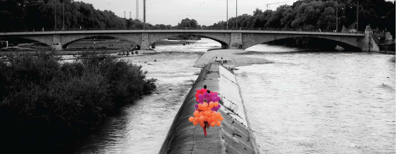 Black and white photograph of the Cornelius Bridge looking south towards the Reichenbach Bridge. On a concrete embankment of the River Isar, known as the Wehrbrücke, three women are walking, each wearing a balloon costume. One woman is wearing a costume made of orange balloons, the second is wearing a costume made of purple balloons and the third is wearing a costume made of magenta balloons.