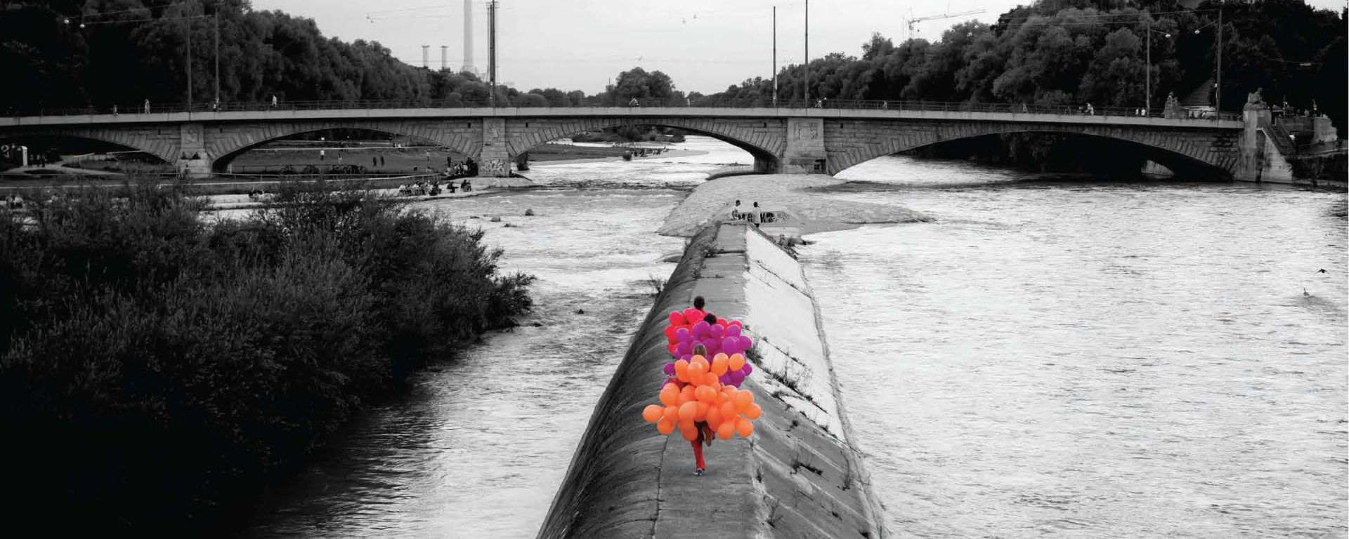 Black and white photograph of the Cornelius Bridge looking south towards the Reichenbach Bridge. On a concrete embankment of the River Isar, known as the Wehrbrücke, three women are walking, each wearing a balloon costume. One woman is wearing a costume made of orange balloons, the second is wearing a costume made of purple balloons and the third is wearing a costume made of magenta balloons.
