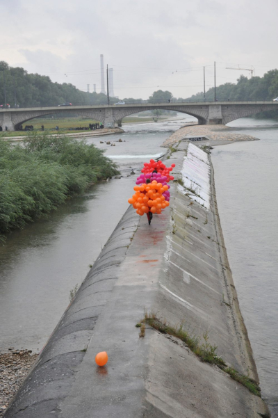 Photograph of the Cornelius Bridge looking south towards the Reichenbach Bridge. On a concrete embankment of the River Isar, known as the Wehrbrücke, three women are walking, each wearing a balloon costume. One woman is wearing a costume made of orange balloons, the second is wearing a costume made of purple balloons and the third is wearing a costume made of magenta balloons.