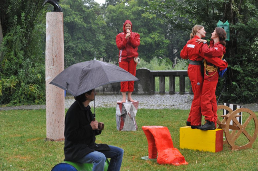 Photograph of a performance on the Isar balcony at the Cornelius bridge in bad weather. There are three performers in red overalls and a spectator holding an umbrella. On the lawn of the Isar balcony are various objects resembling platforms in different shapes, e.g. a platform in the shape of a red shoe, a platform in the shape of a yellow rectangle, and a platform in the shape of a golden wagon wheel. Two performers dance arm in arm on the yellow platform, while the third performer stands on another platform and speaks into a microphone.