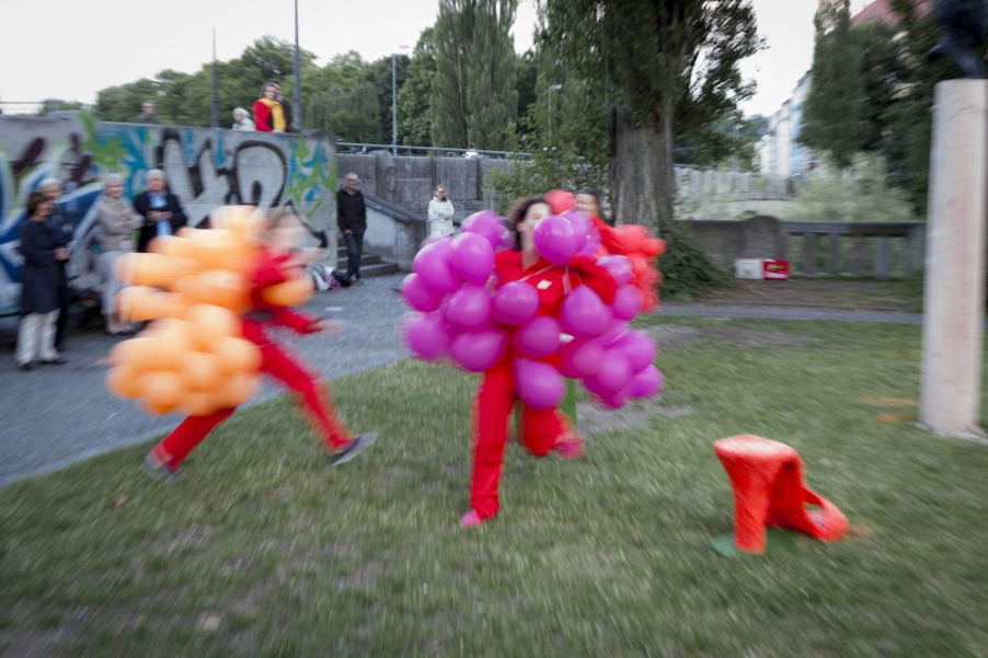 Photograph of a performance on the Isar balcony by the Cornelius Bridge. Three female performers wearing red overalls and balloon costumes in red, orange and purple are running on the lawn. They are surrounded by an audience.
