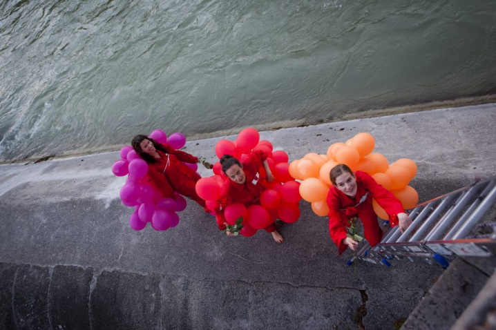 Photograph taken from the Cornelius Bridge of the concrete embankment in the River Isar next to the bridge, known as the Wehrbrücke. Three women are standing on the embankment, each wearing a balloon costume. One woman is wearing a costume made of orange balloons, the second is wearing a costume made of purple balloons and the third is wearing a costume made of magenta balloons. A ladder has been lowered from the bridge onto the rampart and the first woman is about to climb it.