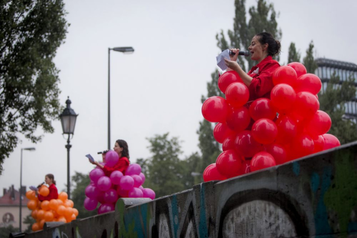 Photograph of three women standing on the parapet of Cornelius Bridge. The three women are wearing red overalls over which are balloon costumes. The first woman is wearing a balloon costume made of red balloons, the second is wearing a costume made of purple balloons and the third is wearing a costume made of magenta balloons. All three women are holding a microphone and reading a text from a piece of paper.