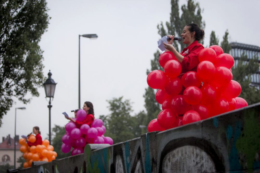 Fotografie von drei Frauen, die an der Brüstung der Corneliusbrücke stehen. Die drei Frauen tragen rote Overalls und darüber Kostüme aus Ballons. Die erste Frau trägt ein Ballonkostüm aus roten Ballons, die zweite aus lilafarbenen und die dritte aus magentafarbenen. Alle drei Frauen halten ein Mikrophon und lesen einen Text von einem Zettel ab.