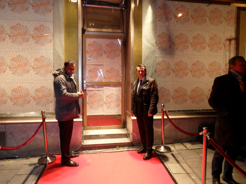 Photograph of the entrance to the building at Blumenstraße 29 at night. The windows of the store and the door on the first floor are covered with A4 posters showing a circular radial graphic with the word "Sanzelize" written on it. A red carpet is rolled out in front of the entrance, flanked by a red velvet cordon. Two men stand at the door like bouncers.