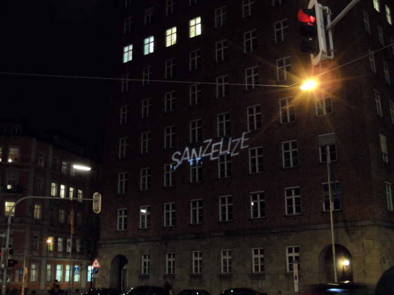 Photo of the multi-storey building at Blumenstr. 29 at night. Four windows on the seventh floor are illuminated. The word "SANZELIZE" is projected onto the façade of the building in capital letters.