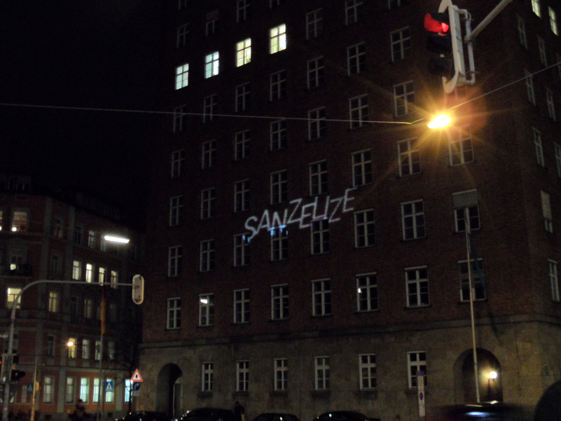 Foto des mehrstöckigen Gebäudes in der Blumenstr. 29 bei Nacht. Im siebten Geschoss sind vier Fenster erleuchtet. Auf die Fassade des Hauses ist in Großbuchstaben das Wort "SANZELIZE" projiziert.