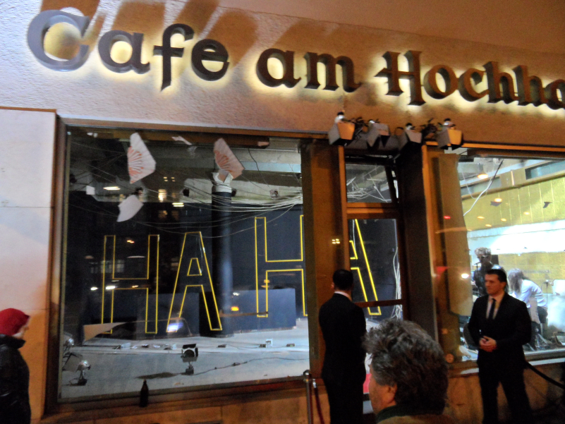 Night-time view of the interior of the building at Blumenstraße 29 through the window front on the ground floor. The words "Cafe am Hochhaus" can be read above the entrance to the shop on the ground floor. Inside the room hangs a yellow neon sign in capital letters that forms the word "HA HA". Two people can also be recognised in the room, one of whom is singing into a microphone. Two men in black suits stand in front of the entrance door, looking like bouncers. The path to the door is flanked by velvet barrier cords.