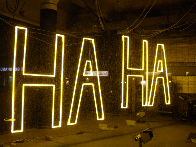 Nächtlicher Blick in das Erdgeschoss des Gebäudes in der Blumenstraße 29 durch ein Fenster. Im Inneren des Raumes hängt ein gelber Neon-Leuchtschriftzug in Großbuchstaben, der das Wort "HA HA" formt.