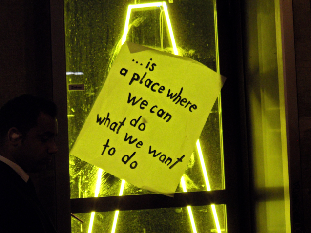 Photography of a window pane at night with a piece of paper stuck at an angle with the following sentence: "... is a place where we can do what we want". Inside the building, the outline of a yellow neon letter can be seen, bathing the room in a green-yellow light.