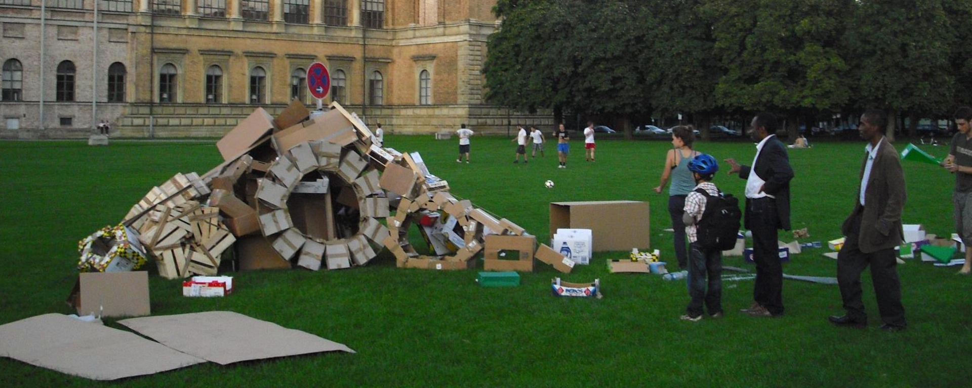 Fotografie einer Installation auf der Wiese hinter der Alten Pinakothek. Die Installation der Künstlerin Valerie Christiansen besteht aus verschiedenen alten Verpackungsmaterialien, vor allem Kartons und Obstkisten aus Sperrholz. Die unterschiedlichen Boxen sind zu Rädern und einer Art Rampe angeordnet, in der Mitte steckt ein Parkverbotsschild. Um die Installation stehen einige Passant*innen, die sie betrachten.