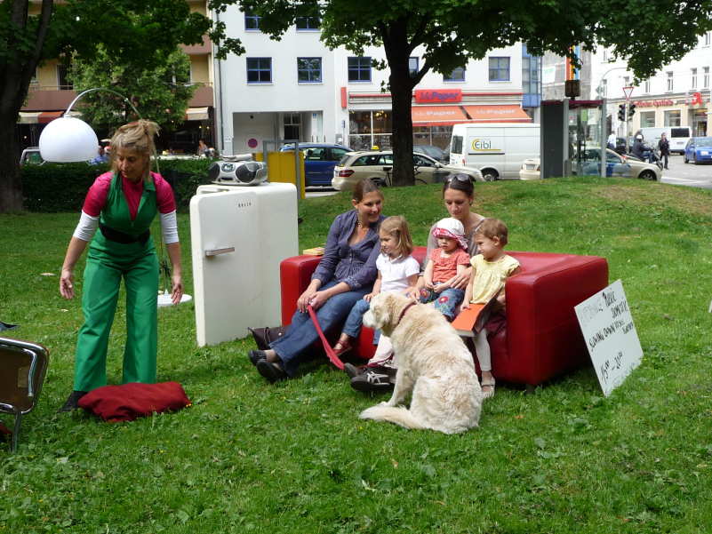 The picture shows a performance on the lawn of Elisabethplatz. Various pieces of furniture are placed on the lawn as if in a living room. An open fridge with a portable CD player on top. Next to it is a pendant lamp with a white shade on one side and a red leather sofa on the other. Two women and three children are sitting on the leather sofa, with a large white dog in front of it. Leaning against the sofa is a sign that reads: "Performance Public Domestic". Slow down to see further 15:00 - 20:00". A performer in a green jumpsuit is moving next to the sofa.