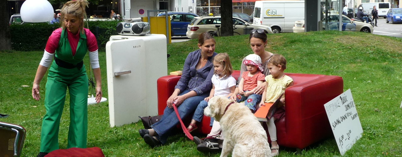 The picture shows a performance on the lawn of Elisabethplatz. Various pieces of furniture are placed on the lawn as if in a living room. An open fridge with a portable CD player on top. Next to it is a pendant lamp with a white shade on one side and a red leather sofa on the other. Two women and three children are sitting on the leather sofa, with a large white dog in front of it. Leaning against the sofa is a sign that reads: "Performance Public Domestic". Slow down to see further 15:00 - 20:00". A performer in a green jumpsuit is moving next to the sofa.