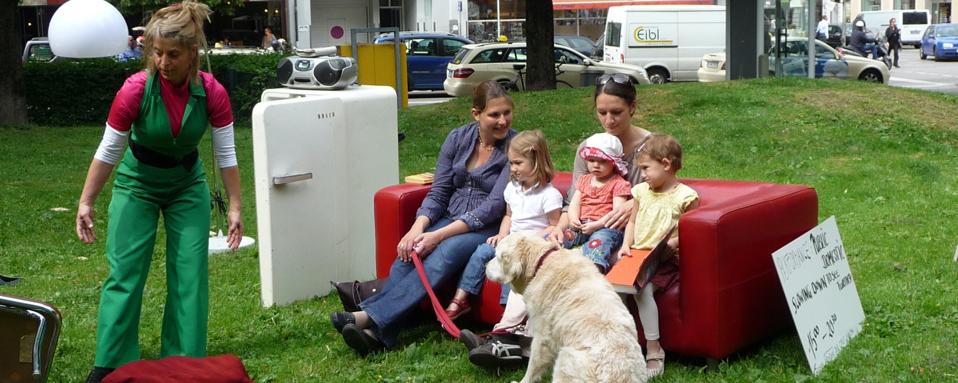 Das Bild zeigt eine Performance auf der Wiese am Elisabethplatz. Wie in einem Wohnraum sind auf der Wiese verschiedene Möbel aufgestellt. Ein offener Kühlschrank, auf dem ein tragbarer CD-Player abgestellt ist. Daneben steht auf der einen Seite eine Pendelleuchte mit weißem Lampenschirm, auf der anderen Seite steht ein rotes Ledersofa. Auf dem Ledersofa sitzen zwei Frauen sowie drei Kinder, davor sitzt ein großer weißer Hund. Gegen das Sofa lehnt ein Schild mit der Aufschrift: "Performance Public Domestic. Slowing down to see further 15:00 – 20:00 Uhr". Neben dem Sofa bewegt sich eine Performerin in grünem Overall.