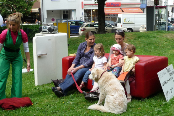 The picture shows a performance on the lawn of Elisabethplatz. Various pieces of furniture are placed on the lawn as if in a living room. An open fridge with a portable CD player on top. Next to it is a pendant lamp with a white shade on one side and a red leather sofa on the other. Two women and three children are sitting on the leather sofa, with a large white dog in front of it. Leaning against the sofa is a sign that reads: "Performance Public Domestic". Slow down to see further 15:00 - 20:00". A performer in a green jumpsuit is moving next to the sofa.