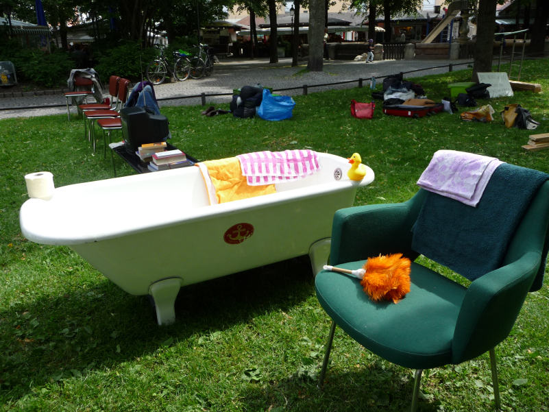 The picture shows a performance on the lawn at Elisabethplatz. Various pieces of furniture are set up on the lawn as if in a living room. In the foreground is a green armchair with a feather duster and two blankets lying on it. Behind it is a white bathtub with a roll of toilet paper, a bath duck and two towels resting on its edge. There are also some brown leather chairs on the lawn and a coffee table on which stacks of books and a television are placed. In the background are several bags, suitcases and boxes containing other household items and textiles.
