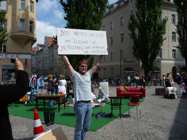 Image of a performance in the square at Münchner Freiheit. A series of furniture and everyday objects are arranged on a green fleece on the ground, giving the installation the appearance of a living room. There is a white fridge, several chairs, a red leather sofa, a table and a clothes rail with clothes on hangers. Performers dressed in white sit in a circle on the floor and appear to be drinking and chatting. In the foreground, a man holds up a white sign that reads: "Please enter and change the positions of the bodies and objects".