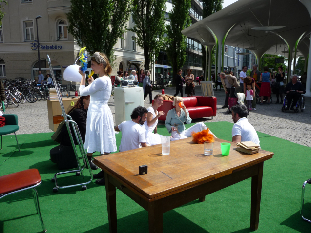 Image of a performance in the square at Münchner Freiheit. A series of furniture and everyday objects are arranged on a green fleece on the ground, giving the installation the appearance of a living room. On display are a chest of drawers, several chairs, a red leather couch and a table. Four performers sit in a circle on the floor, appearing to be drinking and chatting. A fifth performer stands in front of a mirror, combing her hair and holding a hair ornament in her hands. Passers-by can be seen in the background watching the performance.
