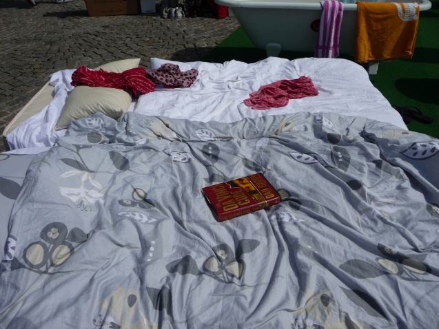 Bild einer Matratze auf dem Plaster auf dem Platz an der Münchner Freiheit. Das Bett mit einem weißen Lacken und einer grauen Bettdecke bedeckt. Zudem liegen darauf verstreut drei Kleidungsstücke sowie das Buch "The Da Vinci Code". Im Hintergrund hinter dem Bett zu erkennen ist eine weiße Badewanne, über deren Rand zwei Handtücher gehängt sind.