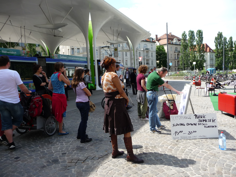 Picture of a small gathering of people in the square at Münchner Freiheit, watching a performance. A white sign names the action: "Performance Public Domestic. Slowing down to see further 15:00 - 20:30. Festival Rodeo of the City of Munich." All that can be seen of the performance are a few structures at the edge of the picture, which look like an arranged living room with a sofa, sofa table and chairs. The 'Münchner Freiheit' bus station can be seen in the background.