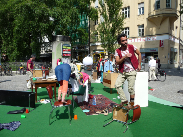 Image of a performance in the square at Münchner Freiheit. A series of furniture and everyday objects are arranged on a green fleece on the floor, giving the installation the appearance of a living room. There is a chest of drawers, a pendant lamp with a white shade, a wooden table, a Persian carpet, a mirror, a clothes rail with clothes on hangers and an overturned coffee table. In between, crockery, books and clothes are scattered chaotically on the floor. Two women and a man, the performers of the work, stand and kneel on three chairs in the middle of this arrangement.