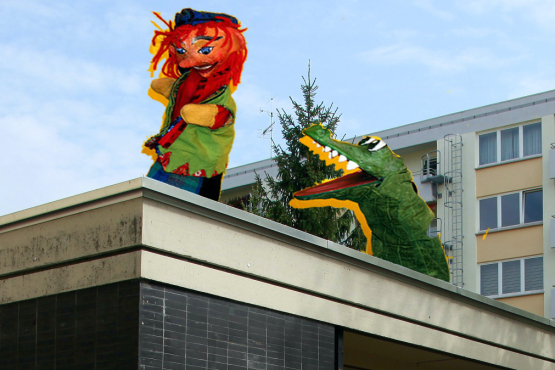 The picture shows a photo collage. A single-storey building can be seen in the foreground and a multi-storey apartment block in the background. A punch and a hand puppet crocodile figure are retouched onto the roof of the single-storey house, performing a kind of Punch-and-Judy-show on the roof.