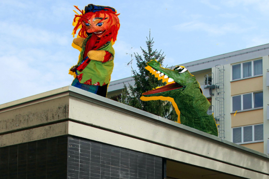 Das Bild zeigt eine Fotocollage. Im Vordergrund ist ein einstöckiges Gebäude zu sehen, im Hintergrund ein mehrstöckiges Mehrfamilienhaus. Auf das Dach des einstöckigen Hauses sind ein Kasperl sowie ein Handpuppen-Krokodilfigur retuschiert, die eine Art Kasperltheater auf dem Dach aufführen.