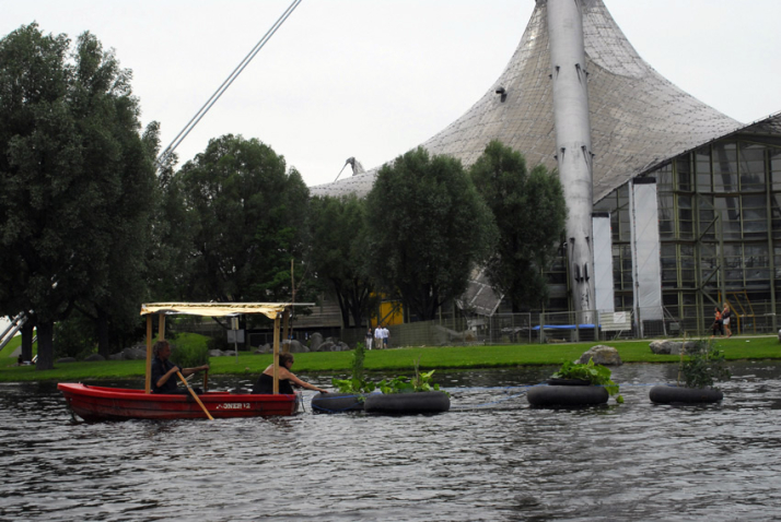 Photograph of a red rowing boat on the Olympic lake. A woman and a man are sitting on the boat under a canopy attached to the boat. Behind the boat there are four black floating rings on the lake, planted with green plants. The woman on the boat is trying to grab one of the rings. The Olympic swimming centre is in the background.