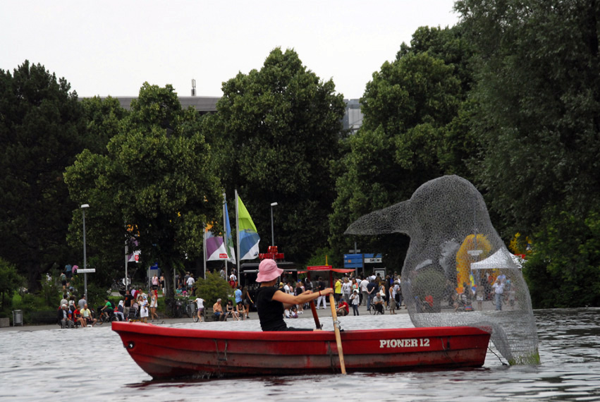 Photograph of a red rowing boat on the Olympic lake. The boat is being rowed by a woman wearing a pink hat. On one side of the boat is a larger-than-life bird made of transparent wire mesh, which appears to be sitting on the boat and looking at the rower. In the background, people are standing on the lakeshore watching the activity on the lake.