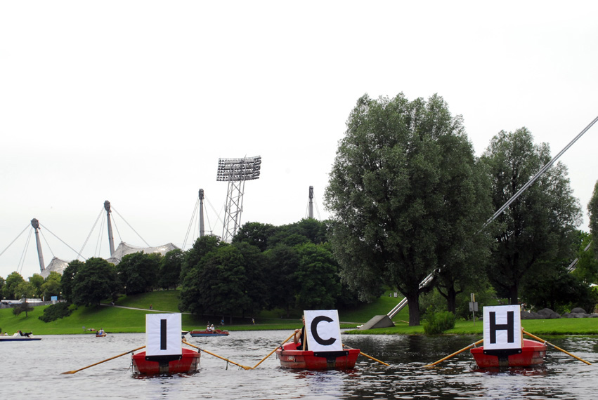 Photograph of three red rowing boats lined up on the Olympic lake. A white placard with a black capital letter is attached to the front of each boat; when read in sequence, they form the word "ICH". The silhouette of the Olympic Stadium can be seen in the background.
