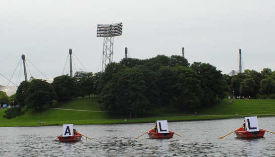 Photograph of three red rowing boats lined up on the Olympic lake. A white placard with a black capital letter is attached to the front of each boat; when read in sequence, they form the word "ALL". The silhouette of the Olympic Stadium can be seen in the background.